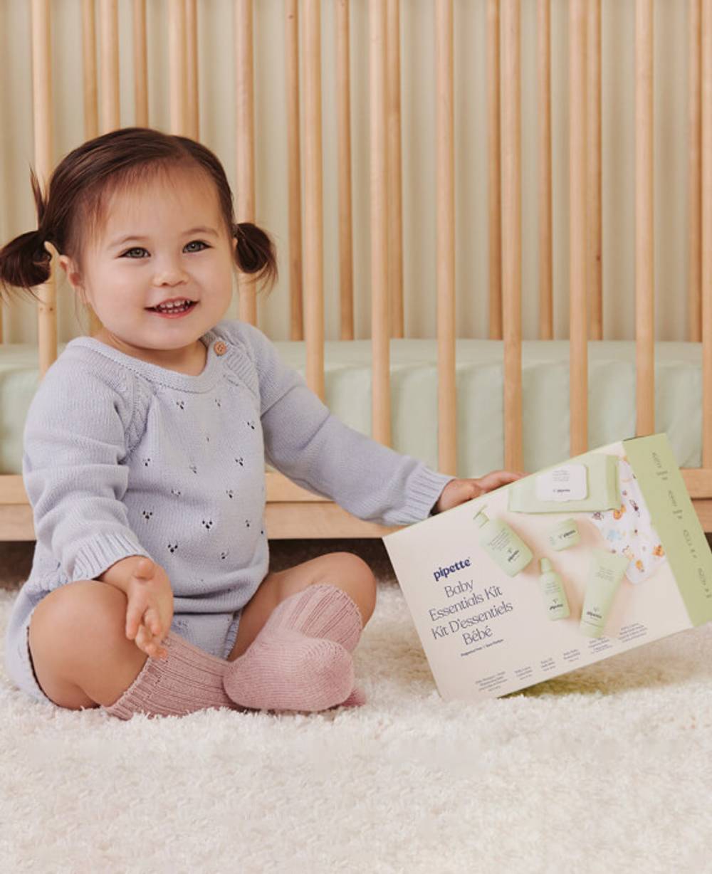 Baby playing with the Pipette Baby Essentials Kit which includes: Fragrance Free Baby Shampoo + Wash 11.8 fl oz, Baby Oil 4.5 fl oz, Fragrance Free Baby Lotion 5.7 fl oz, Baby Balm 2 oz, Baby Wipes 72 ct and a 100% cotton wash cloth.